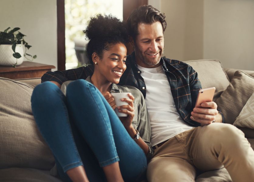 A smiling man and woman sitting on a couch, looking at the phone in the man's hand. 